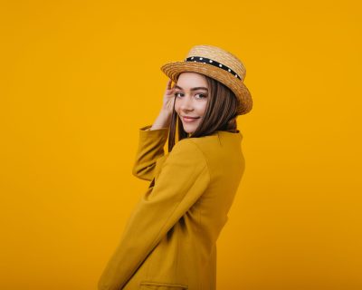Cute young lady in trendy bright jacket posing in studio. Female model in straw hat having fun during photoshoot.