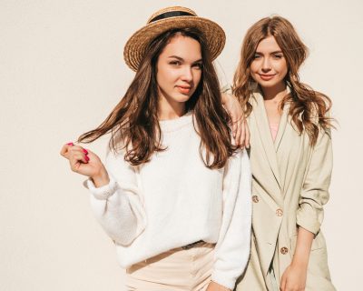 Two young beautiful smiling hipster female in trendy white sweater and coat. Sexy carefree women posing on the street background in hats. Positive models having fun outdoors. Hugging